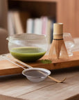 Matcha in een glazen kom met bamboe garde, zeef en lepel op een plank