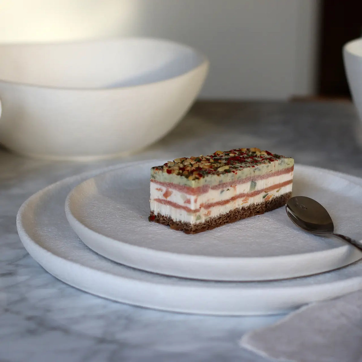 Stuk gelaagde taart op een wit dessertbord met een witte kom erachter