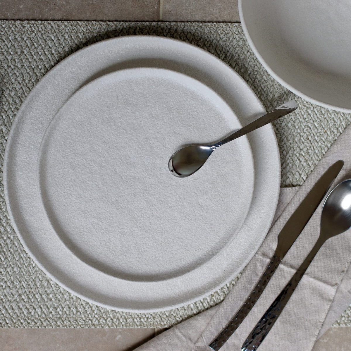 Wit dinerbord met lepel en mes op een geweven placemat, van bovenaf