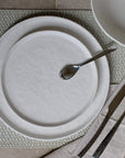 Wit dinerbord met lepel en mes op een geweven placemat, van bovenaf